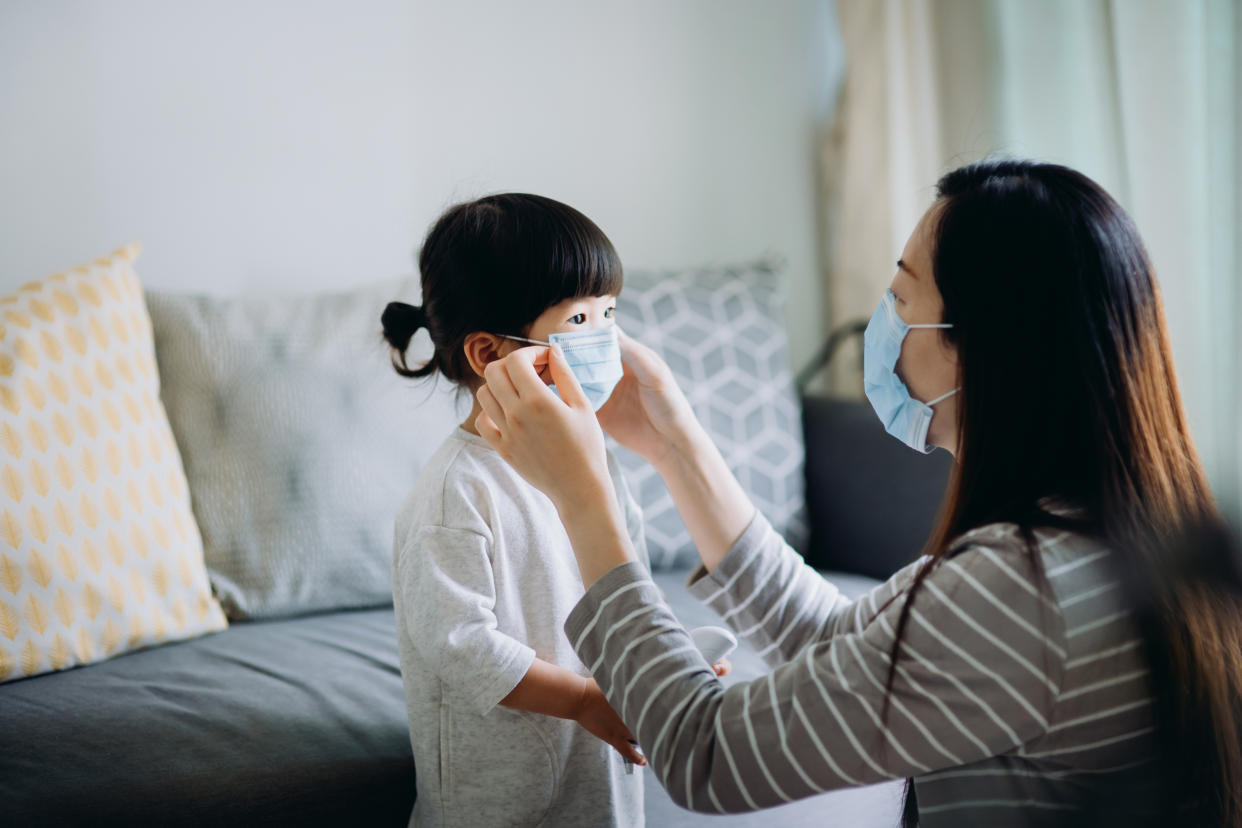 Young Asian mother putting on surgical mask for little daughter before heading out to prevent the spread of cold and flu and viruses