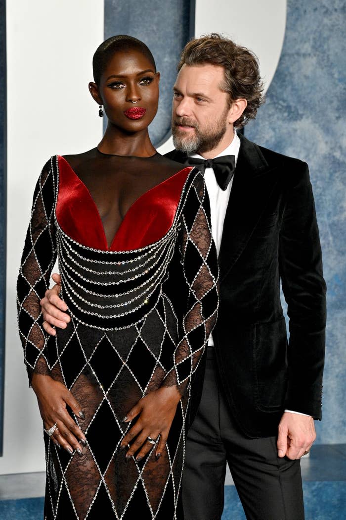 Jodie Turner-Smith in a geometric patterned dress with a deep V-neck and Joshua Jackson in a black suit at a formal event. They pose together closely