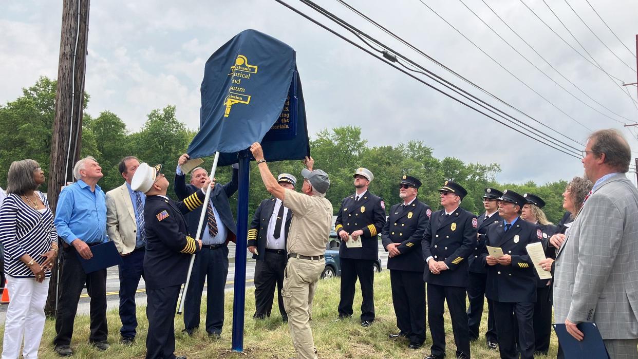 Gene Berry, assistant chief of the Marshalls Creek Fire Company, uses a pole to lift the cover off of a state historical marker for the 1964 Marshalls Creek explosion on June 26, 2023. Holding the cover are Commissioner William V. Lewis Jr., left, from the Pennsylvania Historical and Museum Commission, and Ed Regina, right, who owns Regina Farms, where the marker is located.