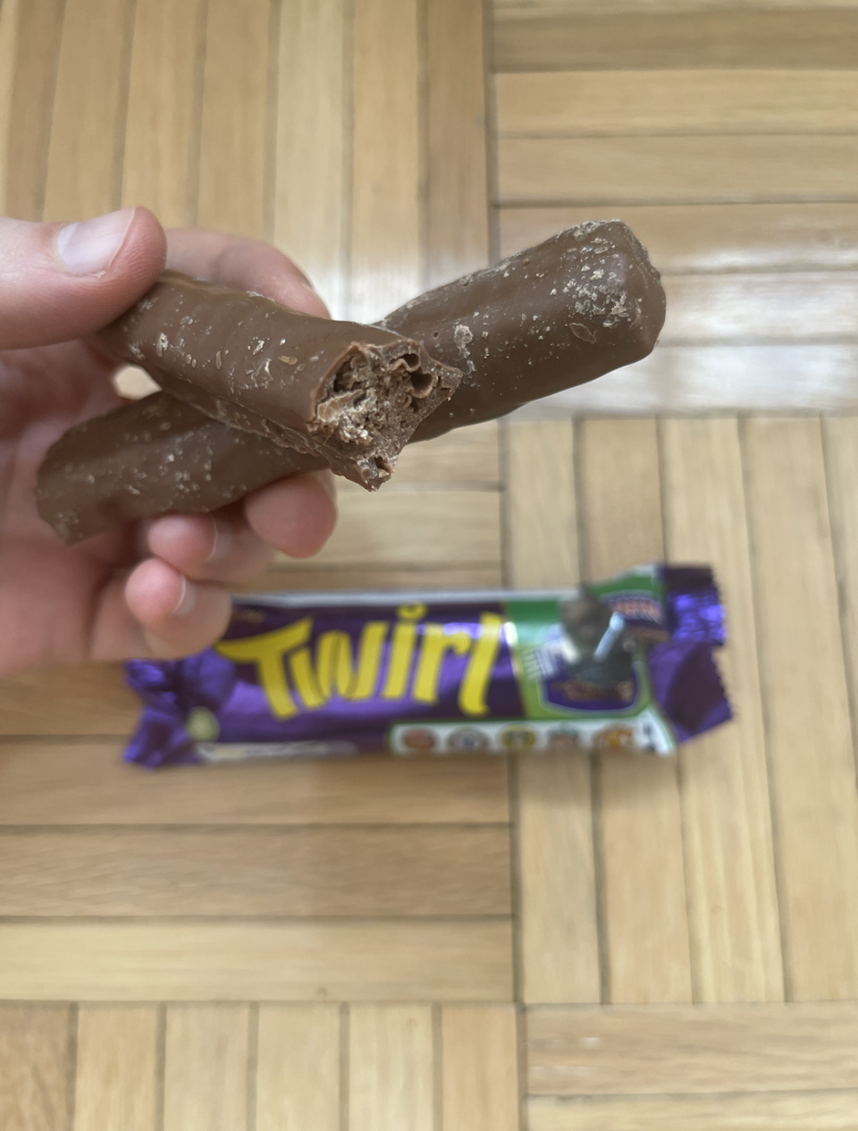 Hand holding a partially eaten Twirl chocolate bar with its wrapper on a wooden floor