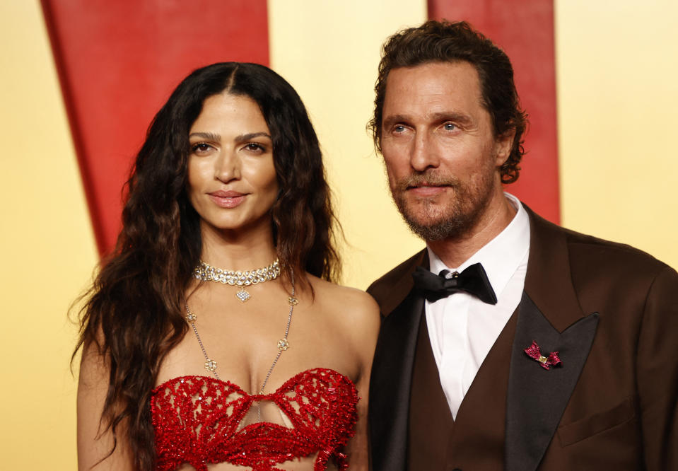 Camila Alves in an ornate strapless dress and Matthew McConaughey in a dark suit with a butterfly pin pose together at an event