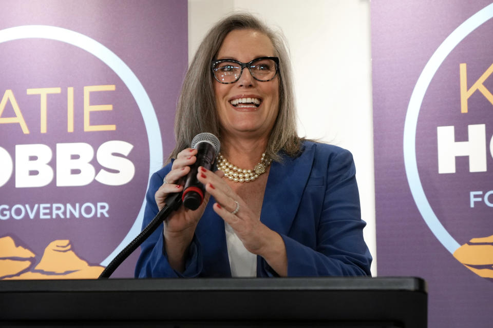 Katie Hobbs at the microphone, looking upbeat, with posters saying Katie Hobbs for Governor behind her.