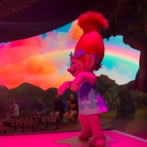 Poppy from Trolls wearing a flower dress and red headband performs on stage with a rainbow background in an animated TVAndMovies scene