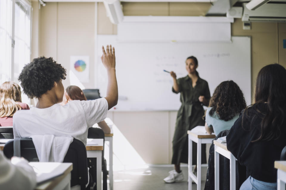 A teacher in a casual dress stands at the front of a classroom, pointing at a student who is raising their hand. Other students are seated at desks, focusing on the teacher