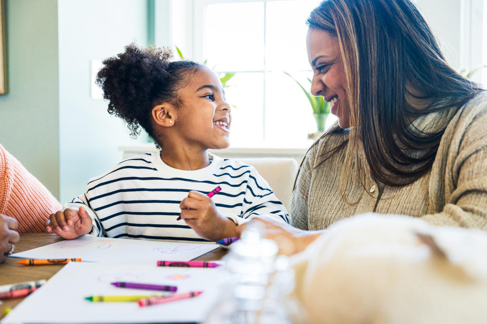 A young Black girl wearing a striped T-shirt and holding a crayon as she draws on paper smiles at her mother