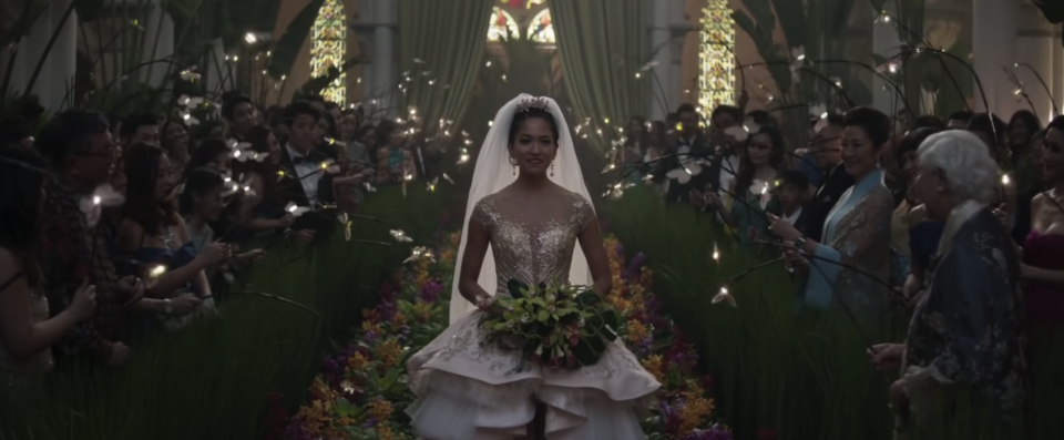 A bride walks down the aisle of a church holding a large bouquet of flowers, guests are holding glowing orbs on sticks