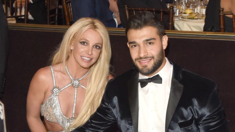 britney spears and sam asghari sit at a dinner table and smile at the camera, she wears a glittering silver dress with cutouts, he wears a black tuxedo and rests one arm across her lap under the table
