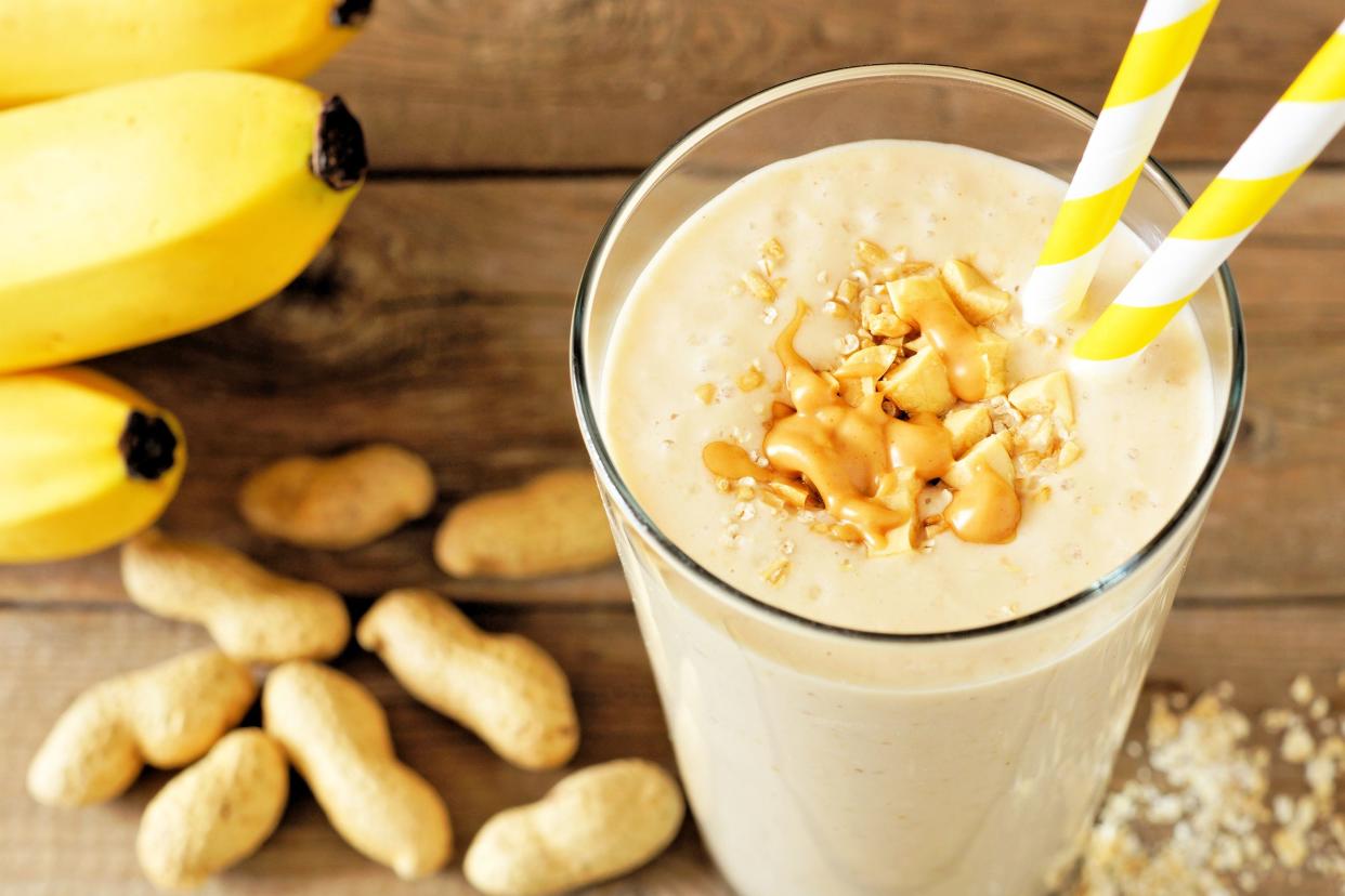 Focus on peanut butter smoothie in a glass with two yellow and white striped straws, surrounded by blurred ingredients on a wooden table