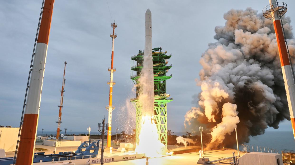  White rocket rising off the launch pad with a green launch tower and gray sky in the background. 