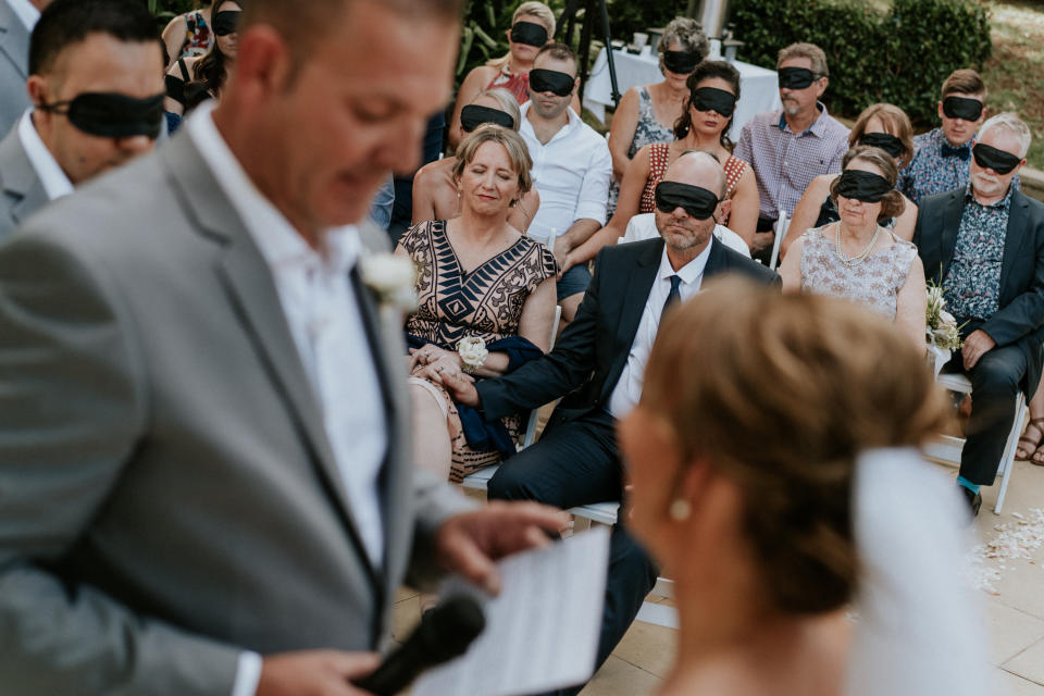 Wedding guests wear black blindfolds during the ceremony. One woman in the center of the shot is not wearing a blindfold: Agnew's mom, who also has cone-rod dystrophy.&nbsp;In the foreground and out of focus, you can see the groom in a gray suit and the bride in a veil with her hair pulled back in a chignon. (Photo: <a href="https://www.jamesday.com.au/" target="_blank">James Day Photography</a>)