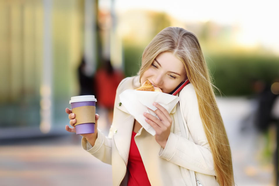 Woman multitasks, holding a coffee cup, talking on a phone, and eating a pastry, outdoors