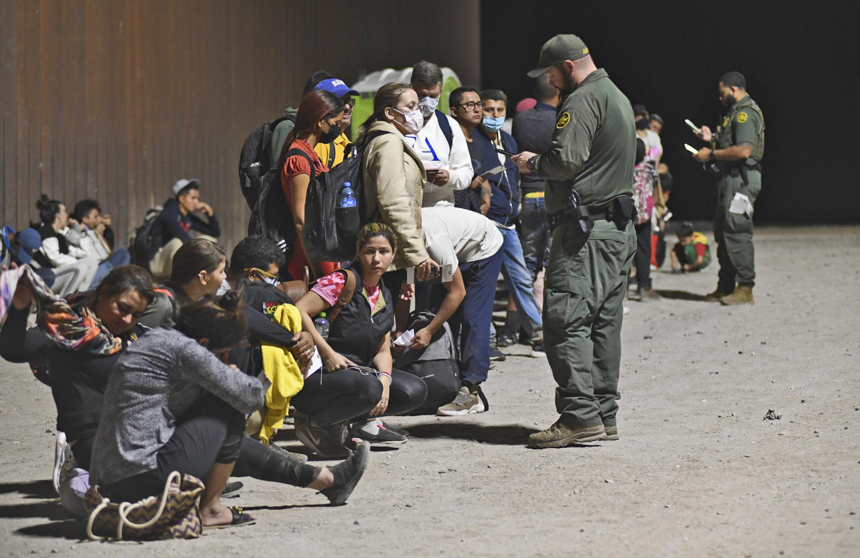 Dozens of migrants sit on the ground or stand as they are detained by several U.S. Customs and Border Protection agents.