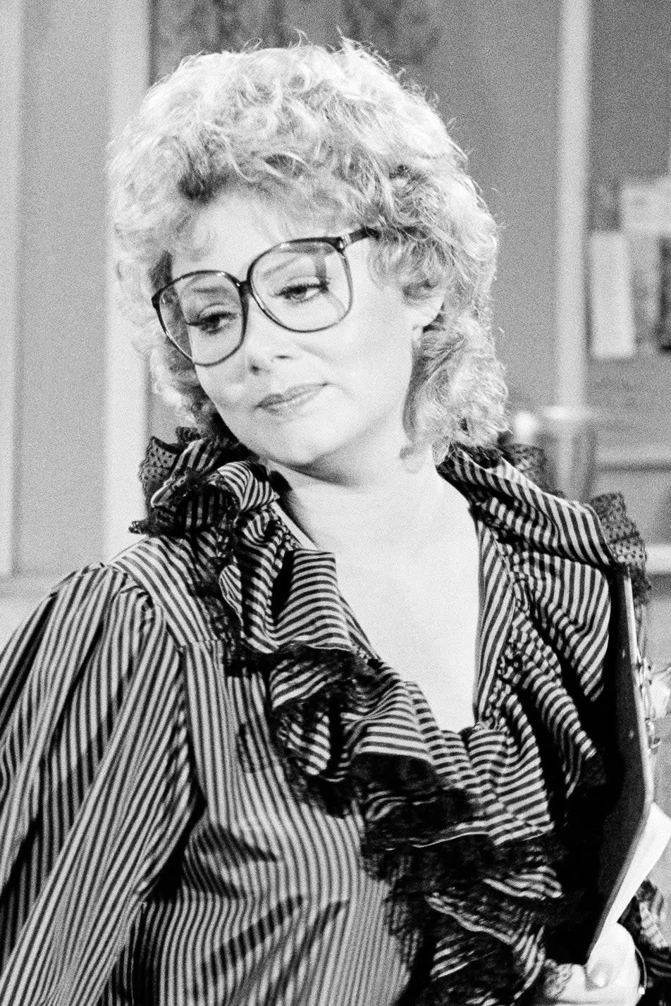 A young Jean Smart in a ruffled blouse and glasses stands in an office setting with two men working in the background. The room has bulletin boards with papers pinned up