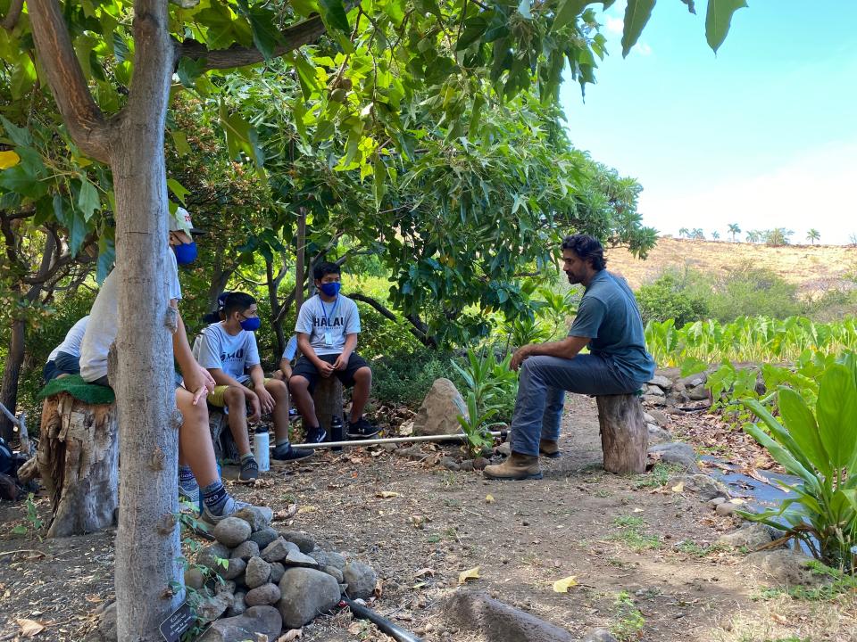 A man sitting on a stump talking to a group surrounded by trees and a field.