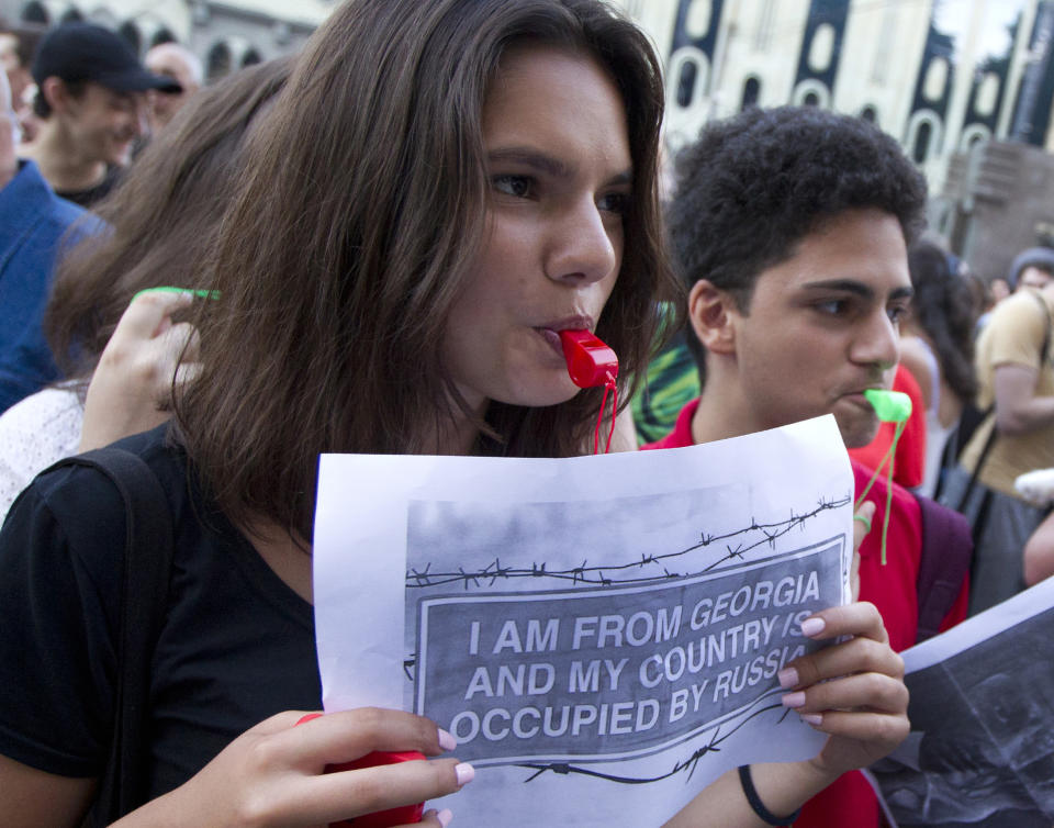 An opposition demonstrator holds an anti-Russia poster as other gather in front of the Georgian Parliament building in Tbilisi, Georgia, Monday, June 24, 2019. Demonstrators have returned to parliament for daily rallies, demanding the release of detained protesters, the ouster of the nation's interior minister and changes in the electoral law to have legislators chosen fully proportionally rather than the current mix of party-list and single-mandate representatives. (AP Photo/Shakh Aivazov)