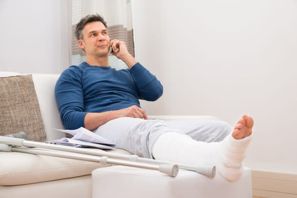 Man talking on phone with broken leg propped up on ottoman while sitting on couch.