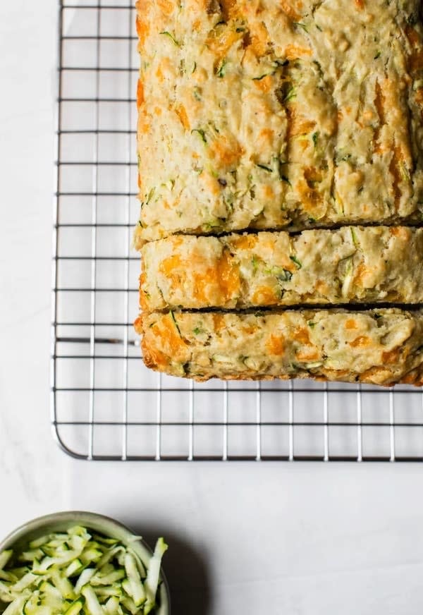 Loaf of zucchini bread on a cooling rack with a bowl of shredded zucchini beside it