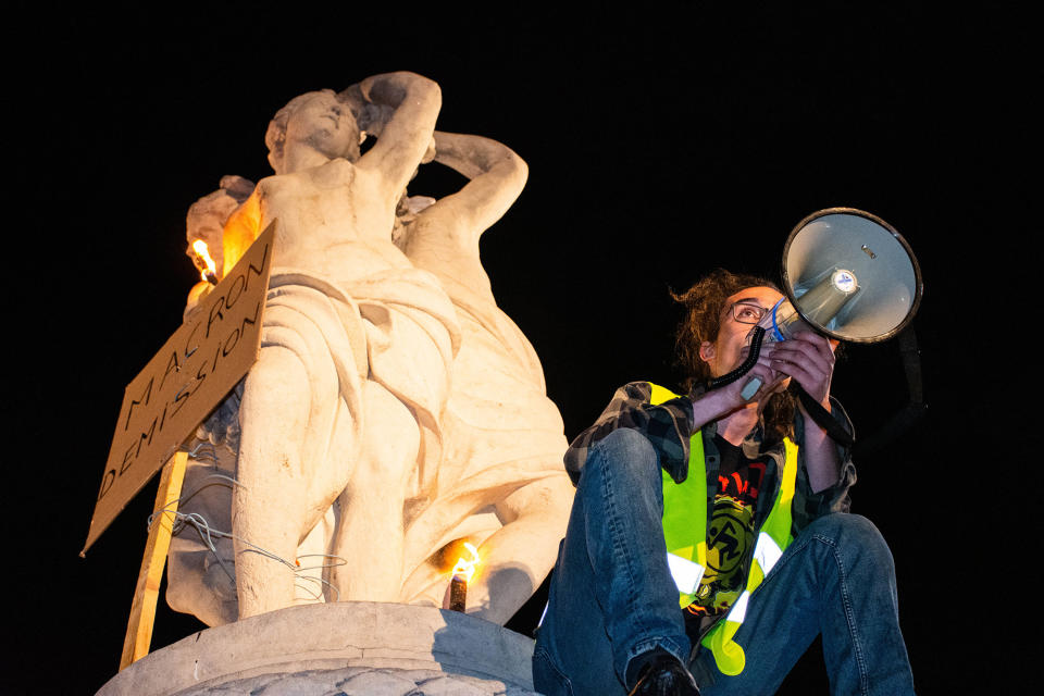 a protester leads the crowd in chants with a bullhorn from the top of a marble statue of three women where a sign says "MACRON DEMISSION" which means "MACRON RESIGN"