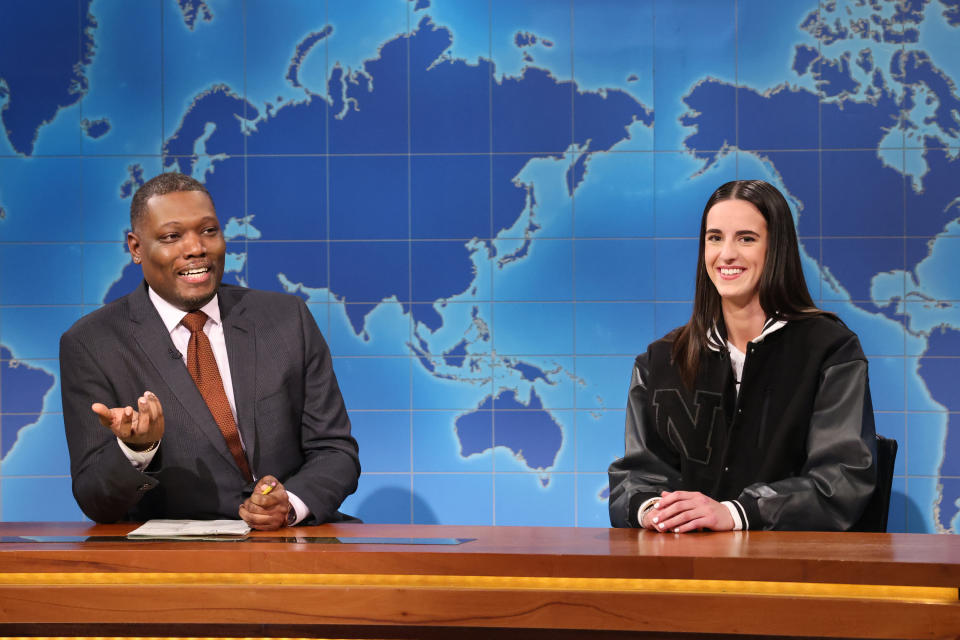 Michael Che and a Caitlin Clark are sitting at the "Weekend Update" desk on the set of Saturday Night Live