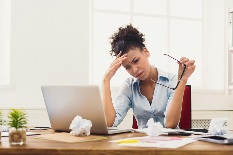 Woman at laptop holding her head; on the table in front of her are crumbled up pieces of paper