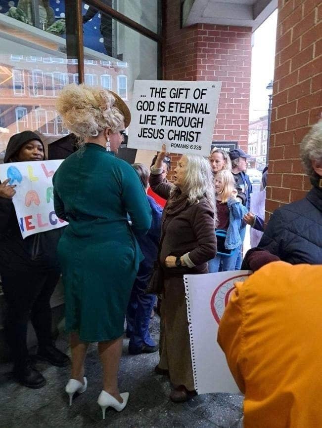 A drag queen wearing white heels and a wig talks to a protester in a group holding a sign reading "the gift of God is eternal life through Jesus Christ"