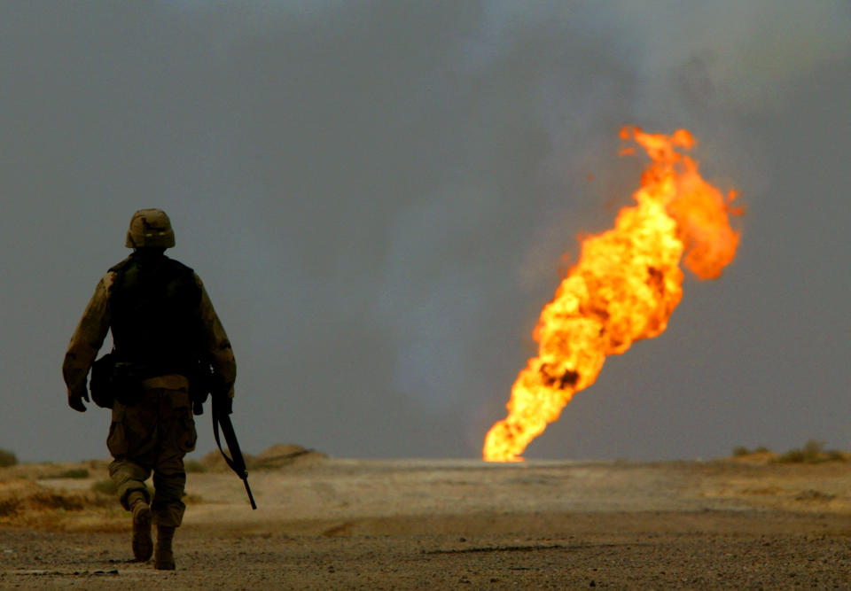 A U.S. Army soldier walks across the desert toward a burning oil well in the distance.