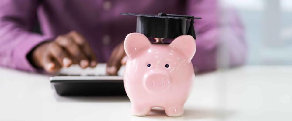 Businessperson's Hand Using Calculator Beside Piggybank With Graduation Hat