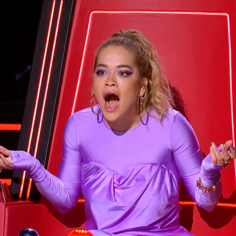 The Voice judge Rita Ora has her mouth open and is visibly shocked, while sitting in The Voice read chair and wearing a lilac outfit. Her hair is in a ponytail and she has hoop earrings on.