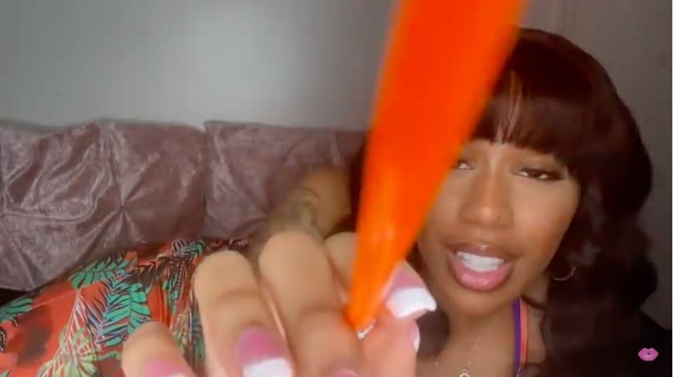 Triniti J ASMR personal attention video. She is pointing at the camera with an orange pen/brush as she lies on the couch