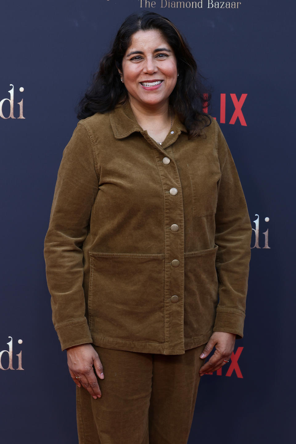 Nisha Ganatra smiles as she poses on the red carpet wearing a brown jacket and matching pants. Netflix and other logos can be seen in the background.