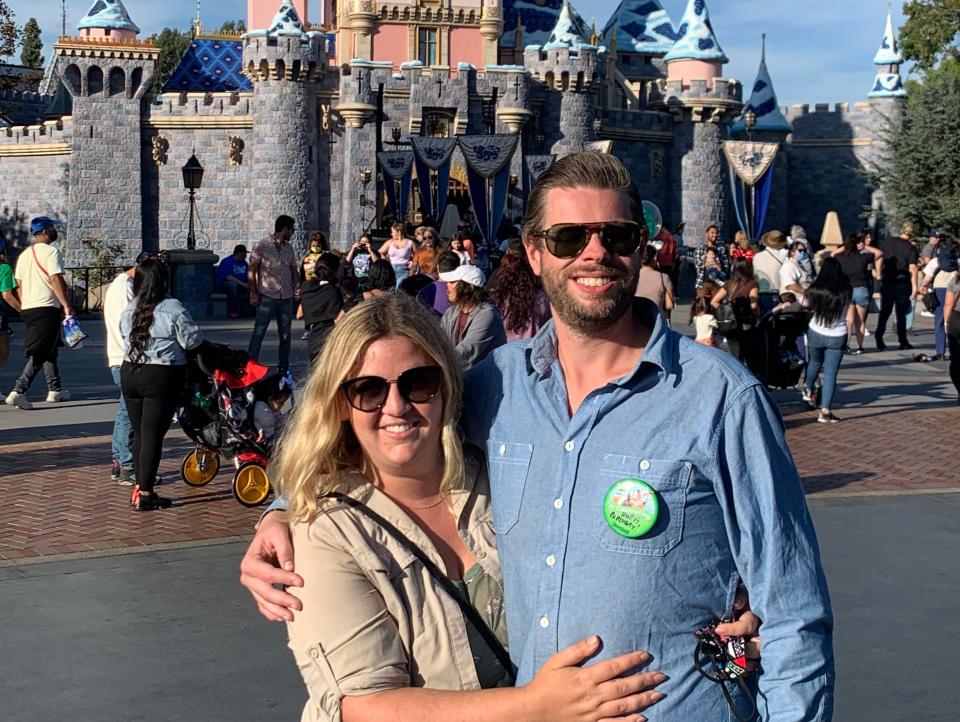 two people posing in front of sleeping beauty castle at disneyland, one wearing a green "i'm celebrating" button