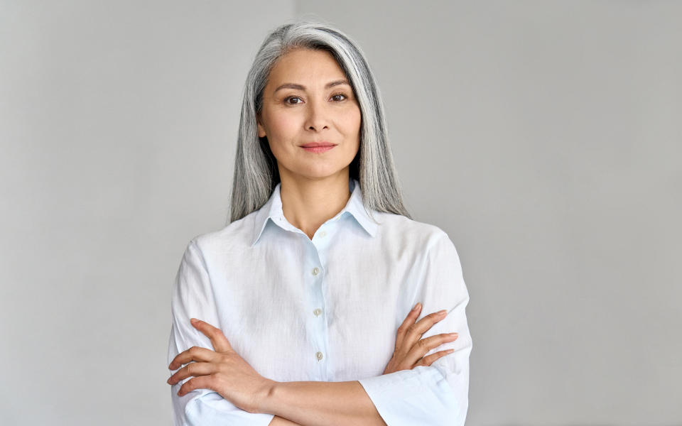 Stylish confident adult 50 year old Asian female psychologist standing with arms crossed looking at camera on gray background.  Portrait of elegant gray hair woman advertising products and services.