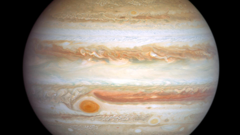     Ein beiger, gasförmiger Jupiter hängt im schwarzen Raum, überlagert wie ein Querschnitt einer geologischen Untersuchung, unten links ein wirbelnder roter Sturm. 