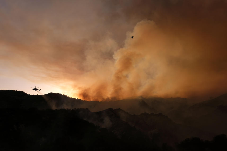 A helicopter drops water as smoke rises from a brush fire scorching at least 100 acres in the Pacific Palisades area of Los Angeles on Saturday, May 15, 2021. (AP Photo/Ringo H.W. Chiu)