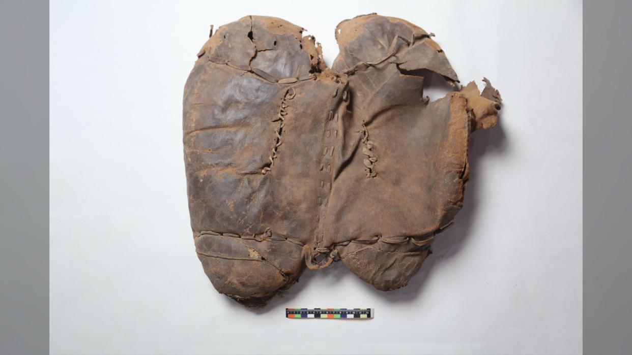  Here we see an ancient leather saddle against a white background. It is brown and kind of heart shaped and looks like a pillow with hand stitching. 