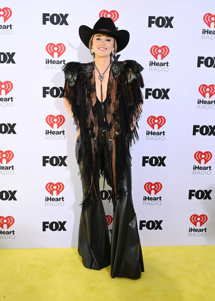 Person on red carpet in a black hat, feathered jacket, and flared pants