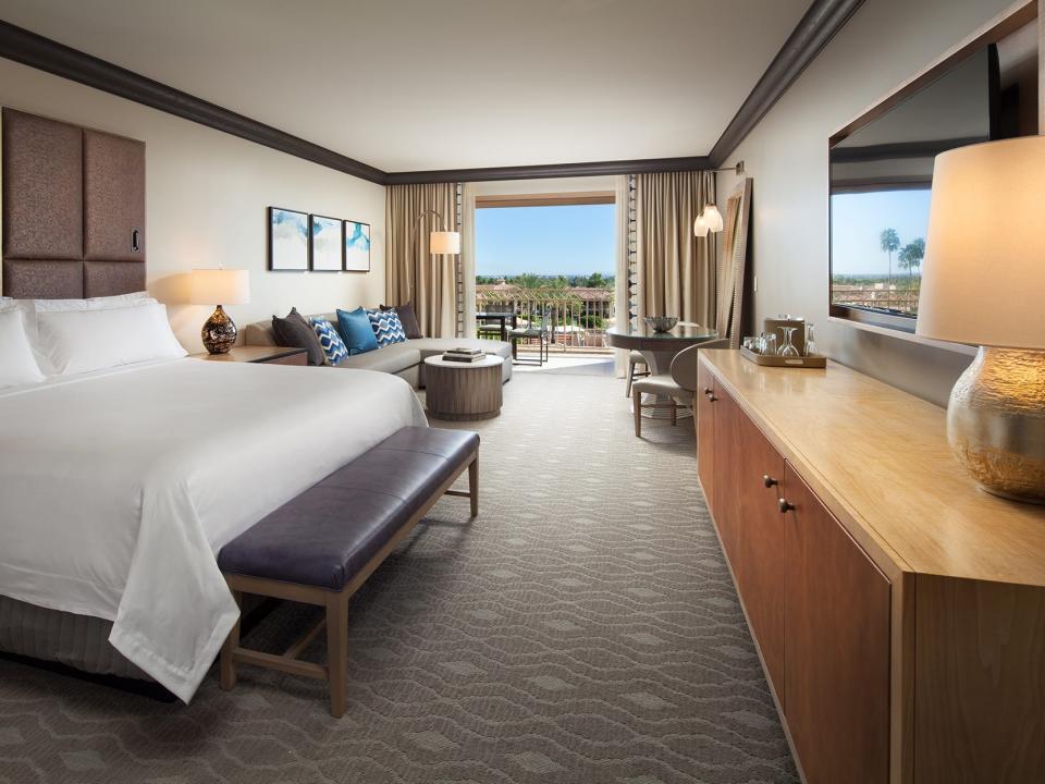 Inside a modern hotel room with a king bed, a selection couch, a sitting area, and a TV and media center. There's a balcony at the back of the room