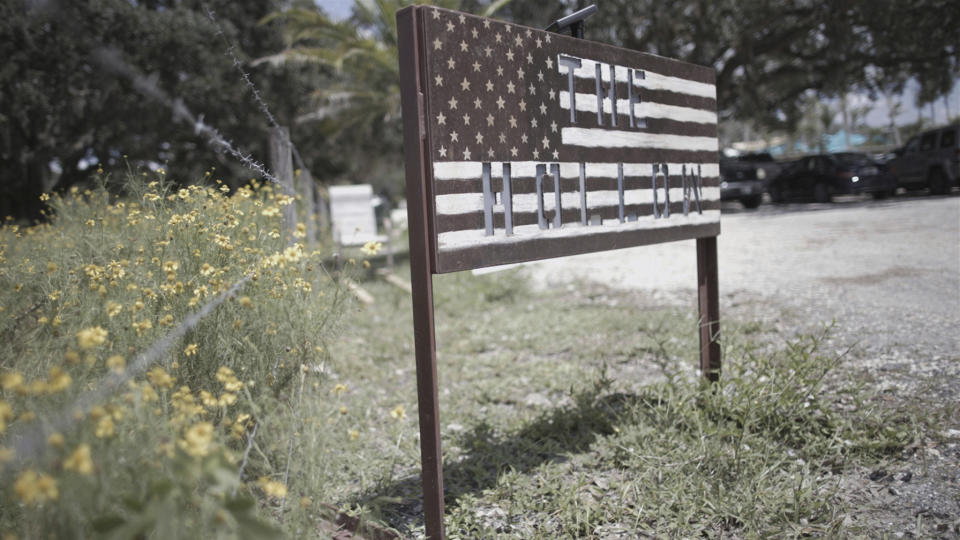 A sign for The Hollow, in the shape of a U.S. flag, stands in a parking lot at the venue in Venice, Fla., on July 31, 2022. After the failed Jan. 6, insurrection, retired three-star general Michael Flynn moved to Sarasota and set out to build a political community of like-minded people. He found an operating base of sorts at The Hollow, a 10-acre site that’s at times a children’s playland, wedding venue, organizing space and weapons training ground. (Richard Rowley/Frontline via AP)