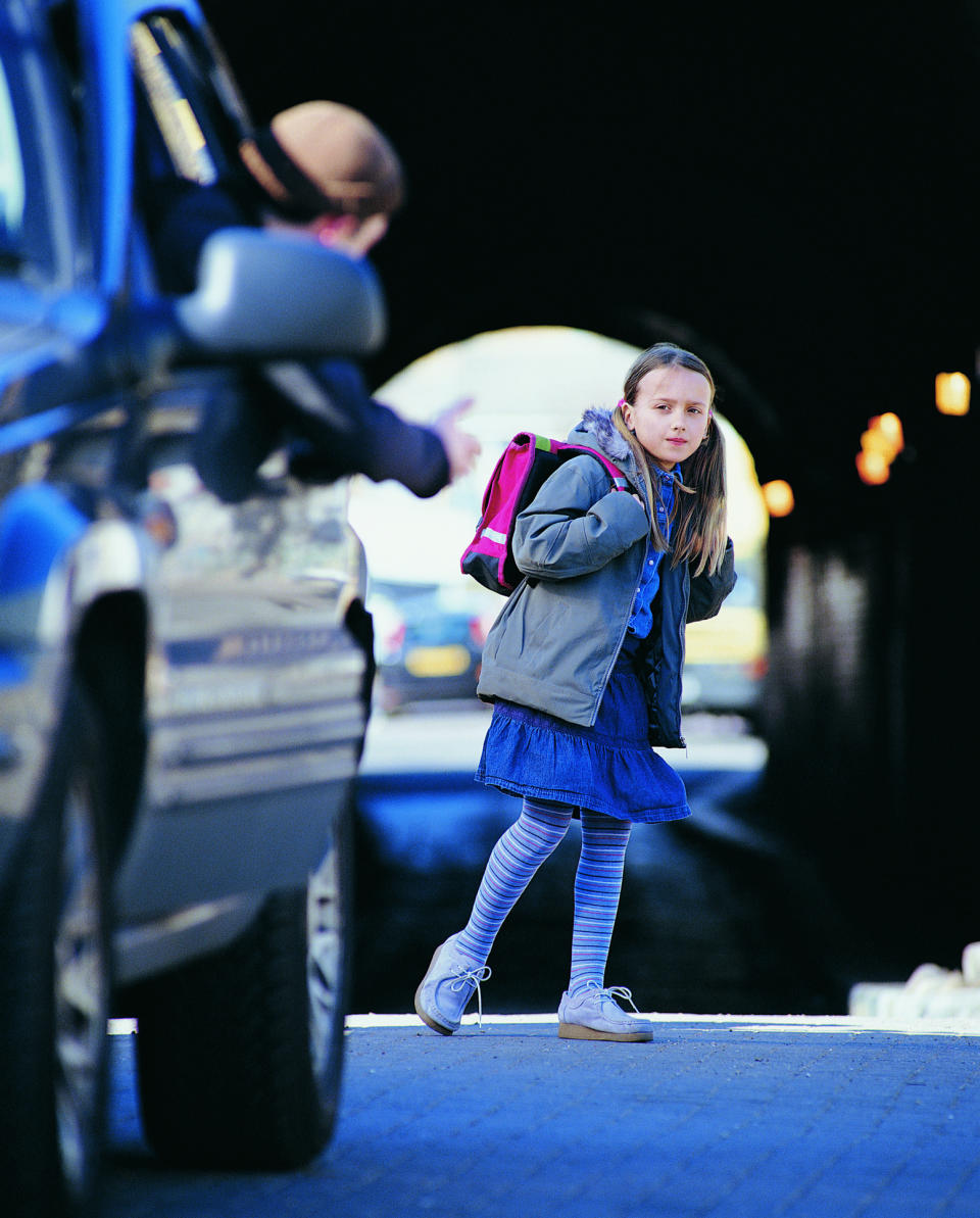 A girl with a pink backpack, wearing a blue dress and striped leggings, looks hesitant near a car with a person gesturing from the driver's seat