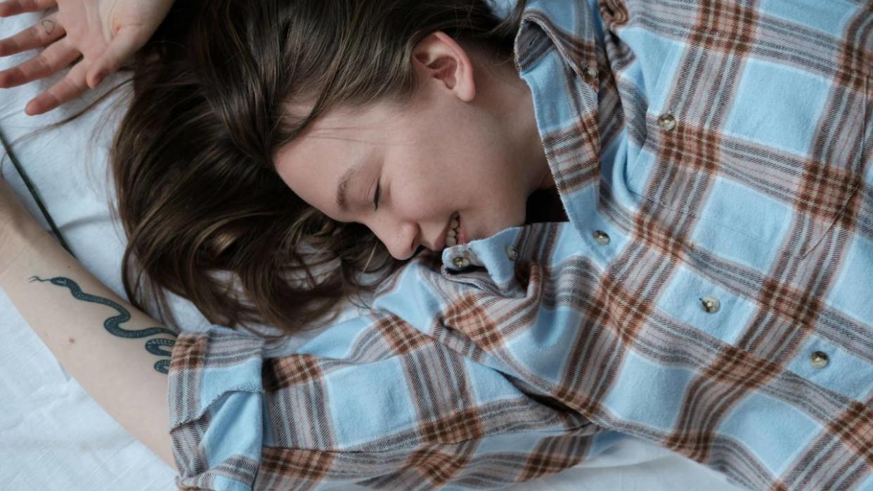  A woman in a checkered shirt, stretching on a bed and smiling while she sleeps. 