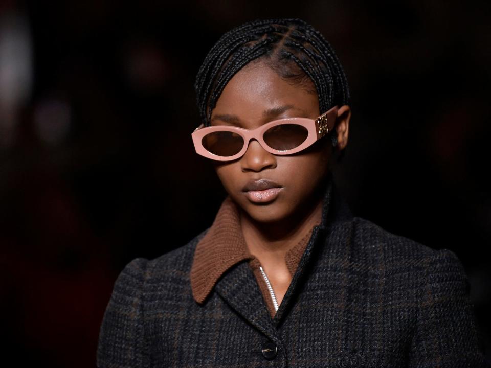 Model wears pink sunglasses, brown sweater, and plaid overcoat during Miu Miu fashion show