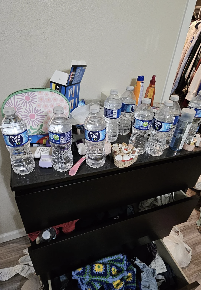 A cluttered dresser with numerous water bottles, some assorted items, and clothing items in the background