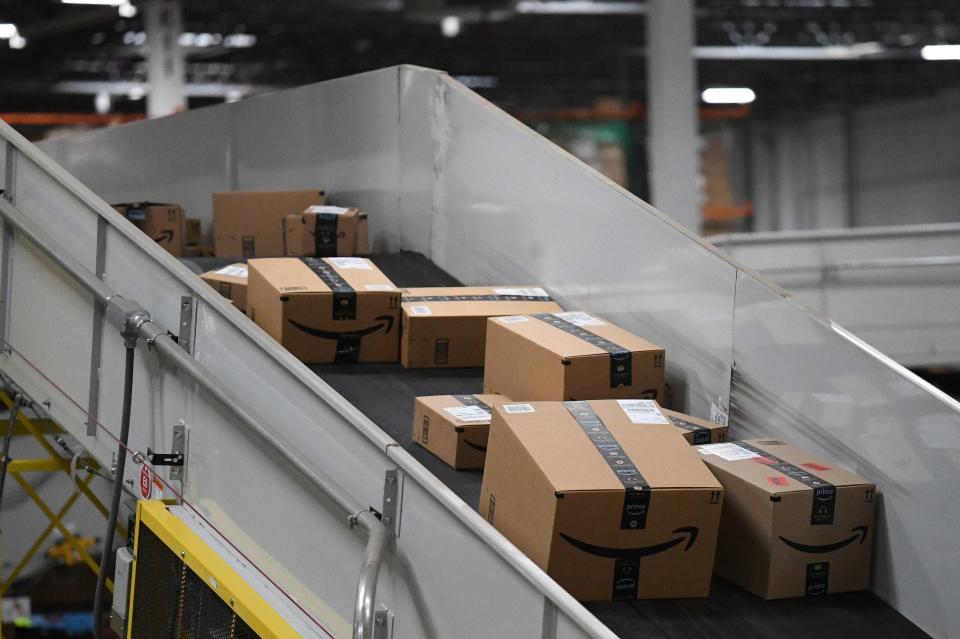 A series of different sized Amazon packages sit on a downwards-sloping conveyer belt with high walls.