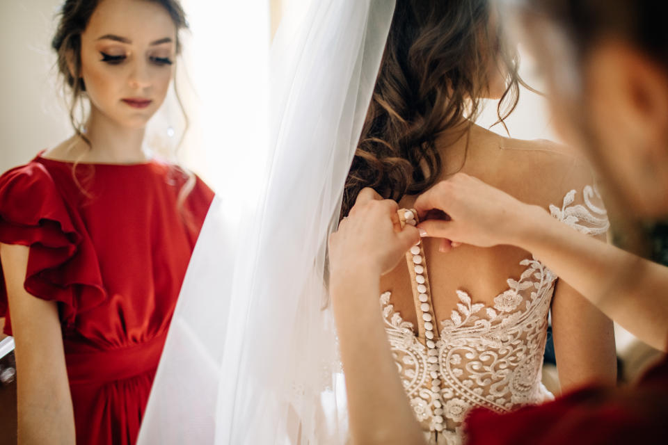 Bridesmaid in a red dress as a man cuttons up back of bride's dress