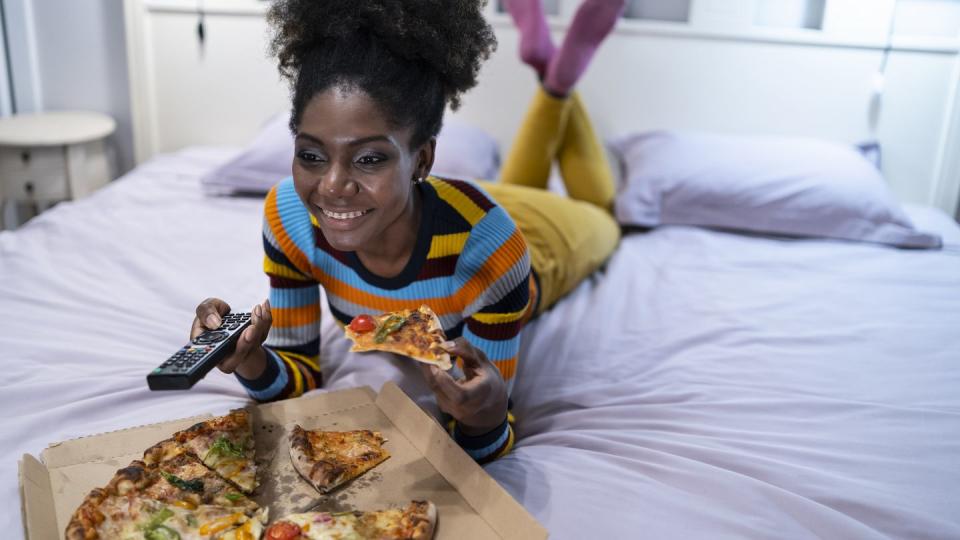 daylight saving sleep - African woman lying down on bed and eating pizza