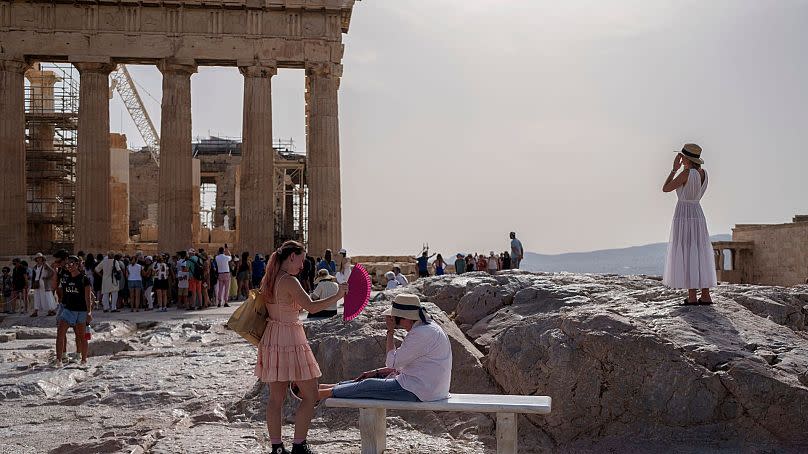 A tourist uses a hand fan to cool down another one sitting on a bench in front of the Parthenon at the ancient Acropolis, in Athens, 12 June 2024. 