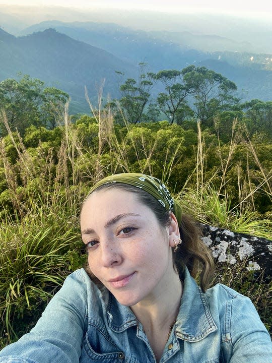 Writer Asia London Palomba is shown in a selfie with the mountains of Munnar, India in the distance. She wears a green headscarf and a denim shirt.