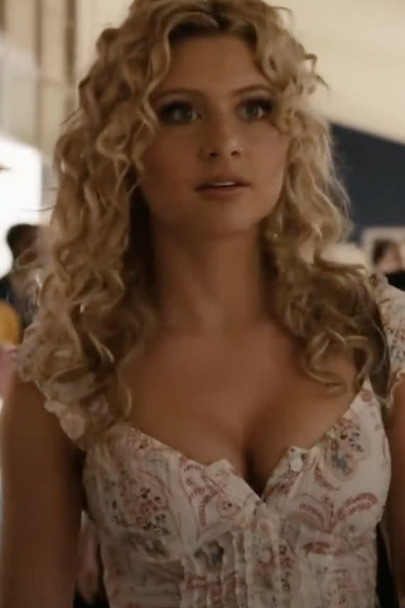 Annie Wilson walking down a school hallway, wearing a patterned blouse, with a concerned expression. Students are visible in the background