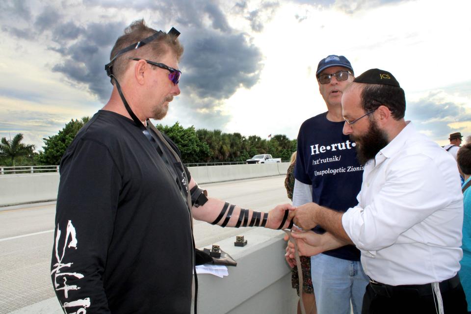 Rabbi Leib Ezagui of the Jewish Community Synagogue wrapped a tefillin, which has leather straps that signify Jewish strength and fortitude in wartime, around the arm of Scott Harris during the rally on the Hood Road overpass to Interstate 95 on Tuesday, Oct. 10, 2023.
