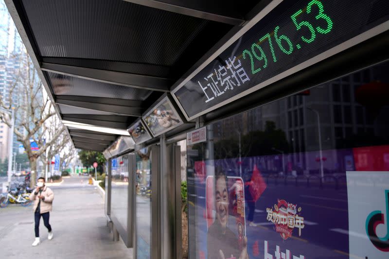 A men wearing a mask walk by an electronic board showing the Shanghai and Shenzhen stock indexes in Shanghai